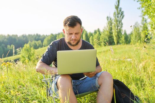 Middle-aged man with laptop in nature. Bearded male sitting on an outdoor chair in meadow in nature, enjoying summer day and using modern technologies for rest and work