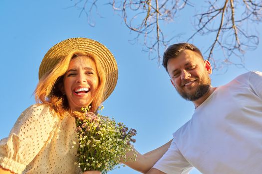 Happy beautiful adult couple looking at camera, blue sky background. Couple in love with bouquet of wildflowers, vacation, relationships, family, lifestyle, leisure, happiness, people concept, looking down at camera
