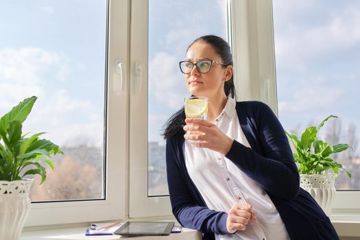 Business woman with glass of water with lemon. Middle-aged female in glasses, white shirt cardigan near window, resting enjoying healthy vitamin water