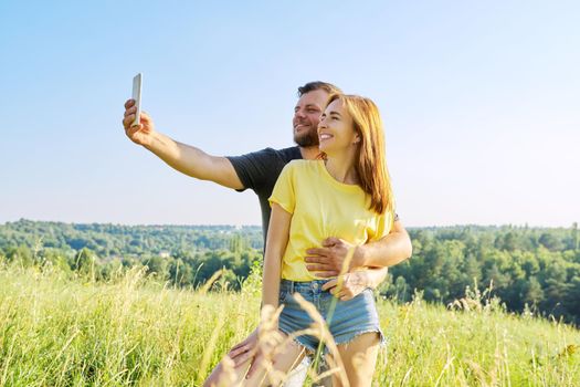 Beautiful happy adult couple taking selfie on smartphone, sunny summer day meadow forest sky background. Relationships, happiness, relaxation, love, people 30s 40s age