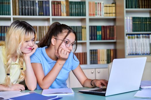 Two girls, students, teenagers 15, 16 years old study in library, using laptop. Education, knowledge, adolescence school concept