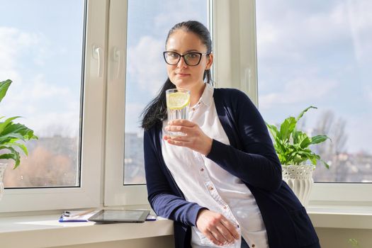 Business woman with glass of water with lemon. Middle-aged female in glasses, white shirt cardigan near window, resting enjoying healthy vitamin water