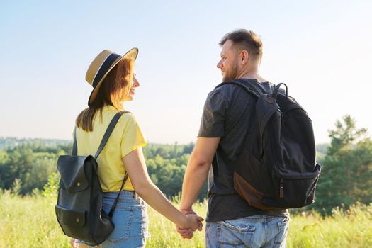 Happy adult couple with backpacks holding hands, back view. Summer nature, sky meadow background, hike, tourism, vacation, relationship, love, lifestyle people concept