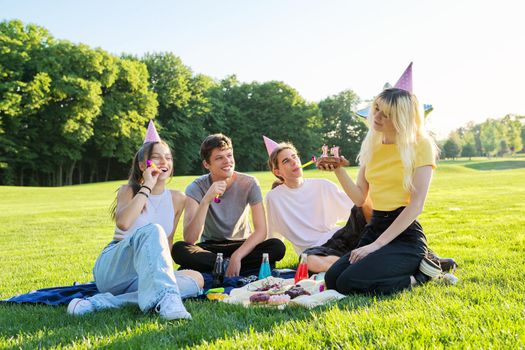 Birthday party. Teenager girl with cake with candles 17, celebrating birthday with friends, teenagers in festive hats sitting on grass in park, summer sunny day