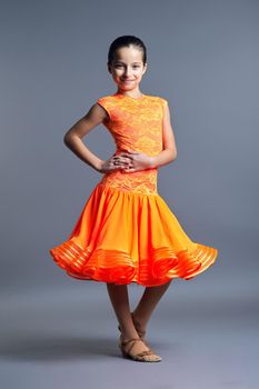 Kids sport dancing, kid girl in orange sport dress posing in dance pose looking at camera on gray studio background.