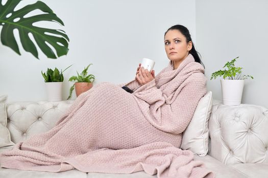 Autumn, winter is a cold season. Woman under a warm knitted blanket sitting on the sofa at home with a cup of hot drink