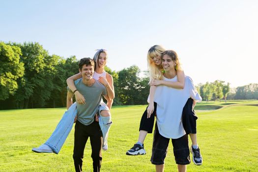 Happy group of teenagers having fun outdoors. Two young laughing couples of teenage friends, outdoors in park, green lawn grass background, sunny summer day. Adolescence, youth, friendship, young people