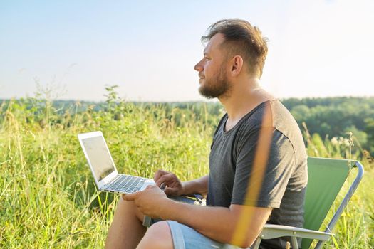 Middle-aged man with laptop in nature. Bearded male sitting on an outdoor chair in meadow in nature, enjoying summer day and using modern technologies for rest and work