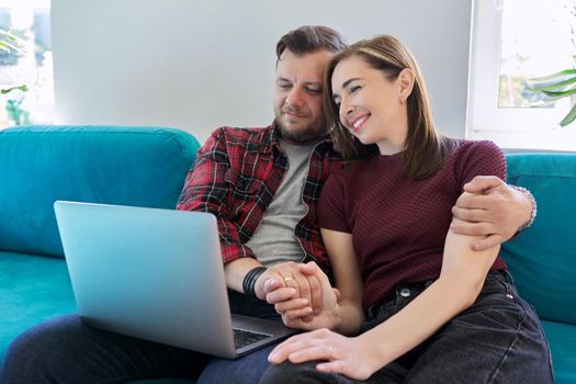 Happy middle aged couple sitting on couch with laptop. Man and woman embracing together looking at laptop screen, in living room. Relationships, lifestyle, communication, home life, middle-aged people