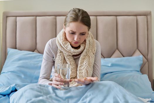 Sick woman with glass of water and pills in her hand, young female sitting at home in bed taking medicine. Medicine, pharmacology, flu season, seasonal diseases, people concept