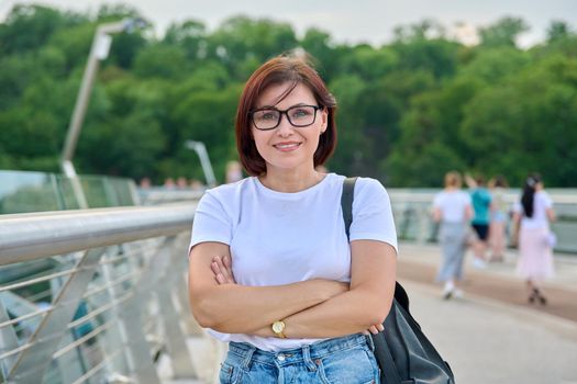 Portrait of smiling confident middle aged woman with crossed arms, summer day in city. Female 40s of age in glasses looking at camera. Lifestyle, urban style, middle-aged people concept
