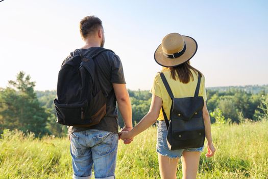 Happy adult couple with backpacks holding hands, back view. Summer nature, sky meadow background, hike, tourism, vacation, relationship, love, lifestyle people concept