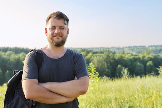 Portrait of confident smiling man in his 40s with folded arms looking in camera. Bearded male with backpack on sunny summer day, sky meadow nature background, copy space