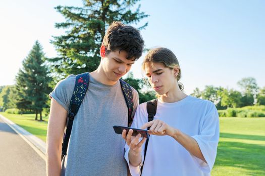 Teenagers friends talking looking at smartphone screen. Two guys 16, 17 years old with smartphone outdoors, on road on sunny summer day. Technology, lifestyle, friendship, youth, adolescence concept