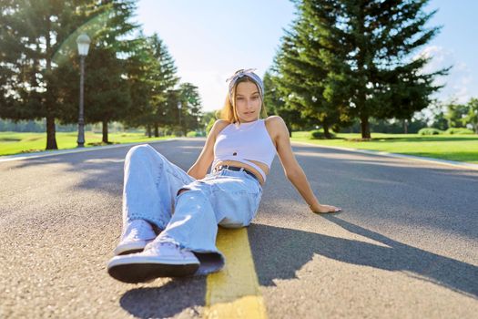 Fashionable trendy teenage hipster female sitting on road in park on sunny summer day. Young stylish model, outdoor. Youth, teens, summer, fashion, street style, lifestyle concept