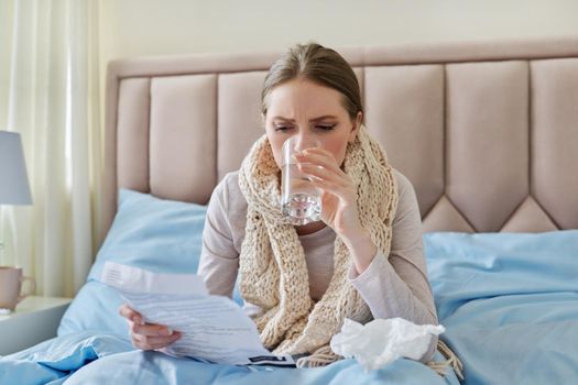 Young sick woman at home in bed, female with glass of water reading instructions and recommendations for medication. Season of colds, viral diseases, health, medicine, pharmacology, people concept.