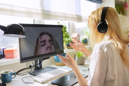 Teen female in headphones talking online with her friend male teenager, using video call on computer, at home at table. Technology, telecommunication, friendship, communication remotely, youth concept