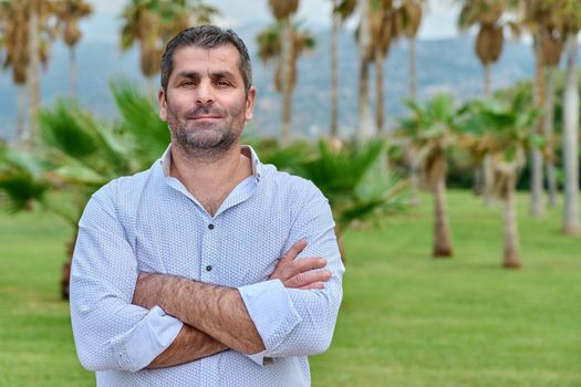 Portrait of confident serious mature man outdoor. Middle aged male looking at camera with crossed arms, tropical park background, copy space. Age, lifestyle, people concept