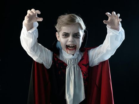 Portrait of boy wearing Halloween vampire makeup and costume cloak bared his teeth, isolated on black background.
