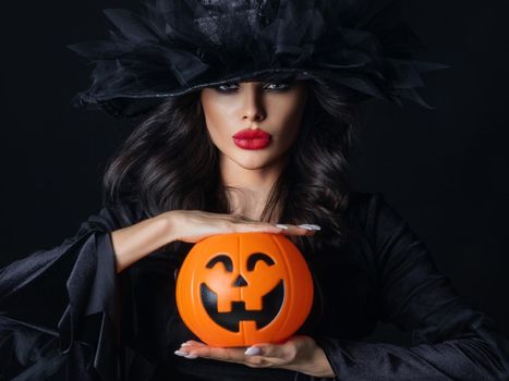 Woman in witch costume hold Halloween pumpkin basket for candies on black background