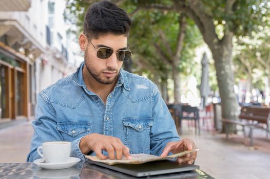 Young traveller man sitting in a cafe terrace and planning her trip with map and laptop. High quality photo
