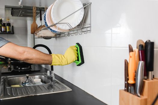 Close up human hand in a yellow rubber glove is using a plastic brush to scrub the tile wall in the kitchen room.