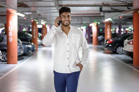 Confident, young handsome man walking along the parking lot while talking on the phone after having parked his car. High quality photo.