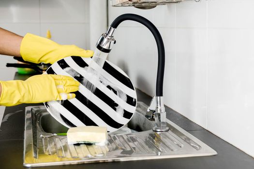 Close up human hands in yellow rubber gloves washing dishes in the kitchen sink.