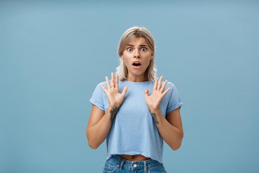 Shocked and worried concerned good-looking feminine woman with blond hair and tattooed arms raising palms gasping and opening mouth expressing empathy and feeling sorry for friend over blue wall. Emotions concept