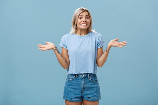 Oops I do not know. Silly cute european girl with fair hair and cool tattoos shrugging with palms spread aside in no idea gesture smiling with sorry look being unaware and clueless over blue wall.