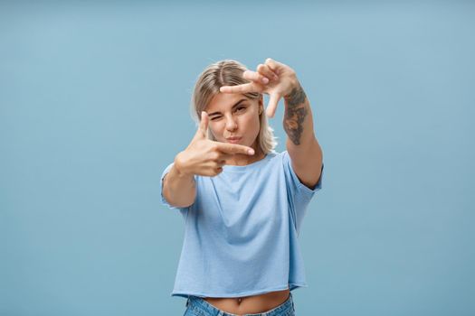 Artistic and creative female designer in stylish t-shirt closing one eye folding lip and making frame gesture while looking through it as if taking measurement or picturing something over blue wall. Copy space