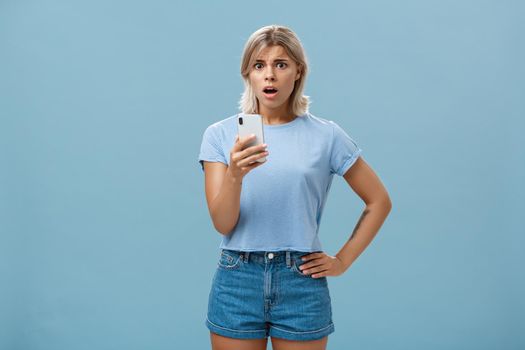 Worried confused young blonde woman in trendy outfit looking concerned and anxious realising bad thing happened receiving shocking news via smartphone opening mouth and staring at camera. Technology concept