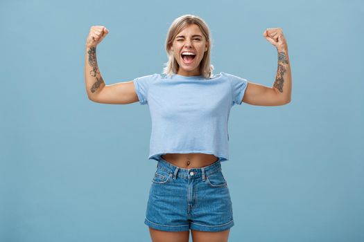 Strong women stand up and fight for rights. Portrait of happy entertained and cool young female athletic blonde with tattoos winking and smiling showing muscles and biceps over blue wall.