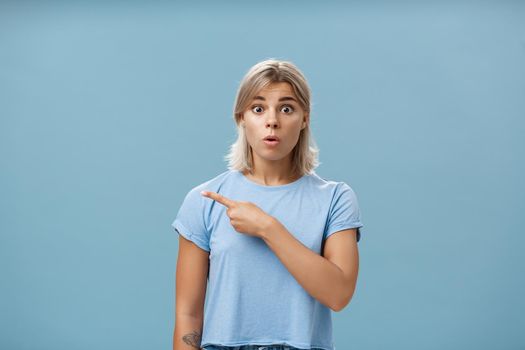 Studio shot of amazed stunned cute blonde witnessing unbelievable event gasping opening mouth staring astonished and pointing left being questioned and shocked over blue background. Emotions concept