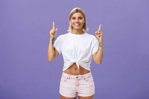 Time look upwards and move forward corporate ladder. Portrait of attractive ambitious and stylish young blonde woman with tattoos and pierced belly pointing up and smiling broadly over purple wall. Copy space