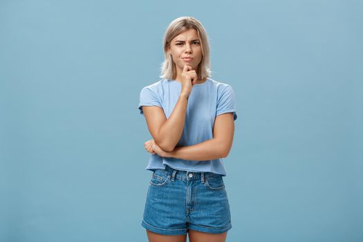 I doubt it good idea. Suspicious intense creative young female coworker in outdoor outfit frowning from doubtful thoughts holding hand on chin while thinking expressing disbelief over blue wall.