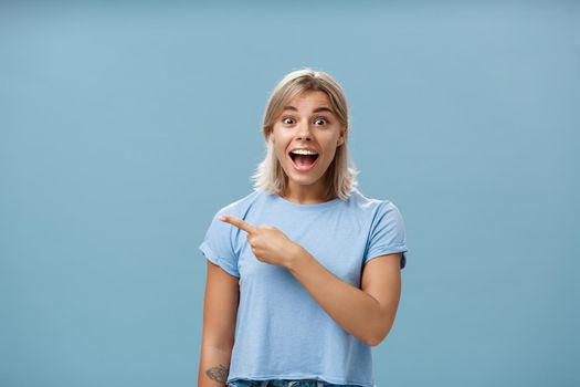 Hurry up and look. Excited and thrilled impressed charming blond female student in casual t-shirt smiling and yelling from amazement and happiness while pointing left staring surprised at camera. Emotions and advertisement concept