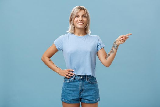 Check it out. Portrait of beautiful confident young caucasian girl with blond hair pointing right and holding hand on waist in self-assured relaxed pose smiling broadly over blue background.