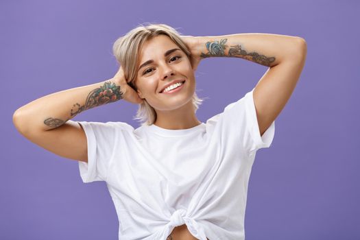 Lifestyle. Close-up shot of good-looking stylish and relaxed blond woman with tattoos on arms holding hands behind head smiling with carefree pleased look enjoying weekends over purple background.