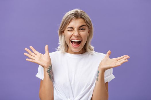 Waist-up shot of playful energized sociable attractive woman with blond hair, tanned skin and tattoos raising hands joyfully and spread aside winking from amusement smiling broadly over purple wall.