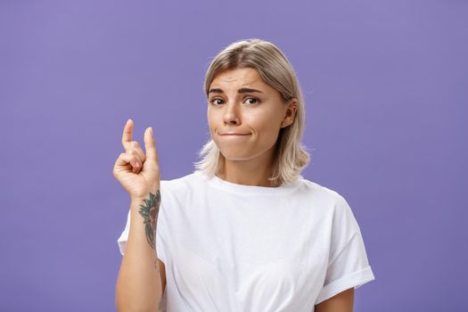 Girl having tiny problem. Concerned attractive blonde girl with tattoo on arm pursing lips in troubled look shaping small or little object, dissatisfied with regret in eyes over purple wall. Copy space