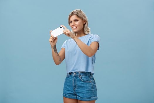 Girl trying to take photo of pet moving fast staring intense at smartphone screen pointing phone camera left feeling focused while taking picture on cellphone over blue background. Technology and lifestyle concept