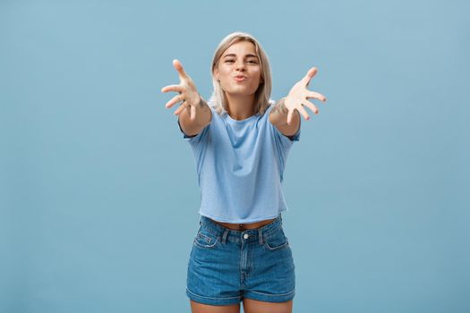 Come to mama. Portrait of friendly joyful and enthusiastic caucasian female student in trendy outfit pulling hands towads camera and folding lips to hug and give passionate kiss smiling over blue wall.