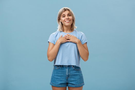 So sweet thanks. Touched beautiful and emotive blond girl in t-shirt and shorts with tanned skin holding palms on breast, sighing and smiling joyfully being thankful and grateful over blue wall. Emotions concept