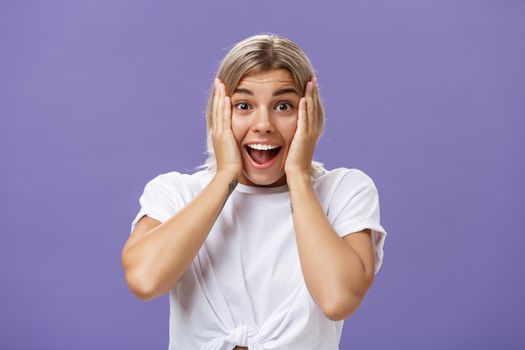 Amazed and thrilled happy european woman with fair hair clean skin and white teeth smiling joyfully holding hands on cheeks being impressed and amused with awesome news over purple wall. Emotions concept