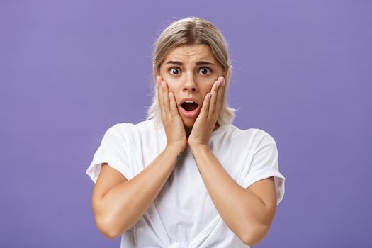 Shocked and worried silly european woman expressing empathy feeling sorry for poor animal in danger gasping opening mouth holding hands on face frowning feeling troubled over purple wall. Emotions concept