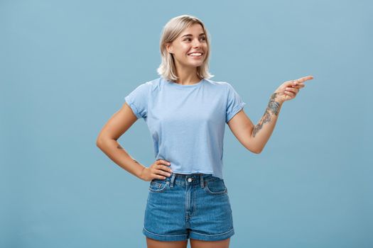 Bossy confident female manager with tattoo on arm holding hand on waist pointing and gazing right with pleased relaxed look giving directions to employees posing over blue background. Copy space