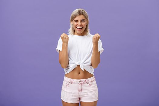 Rejoicing satisfied and pleased happy young ambitious girl with blonde hair in trendy white cropped top and shorts saying yes winking and smiling clenching fists in success or victory sign. Emotions and body language concept