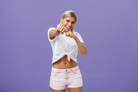 Ready fight back. Energized happy and awesome blonde female with tanned skin and fit body pulling fist in attack gesture towards camera smiling joyful acting like fighting or boxing over purple wall.