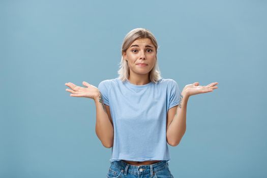 No idea how help sorry. Portrait of unsure gloomy girlfriend feeling regret shrugging with hands near shoulders frowning and pursing lips being uncertain and unaware over blue background. Body language concept